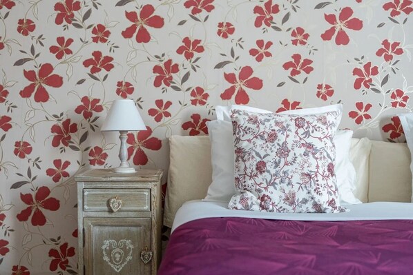 Un dormitorio con papel tapiz floral, una mesita de noche con lámpara, una cama con manta morada y un cojín decorativo.