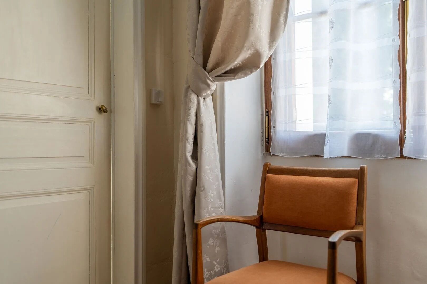 Een kamer met een houten stoel, een witte deur en een raam met dunne gordijnen.