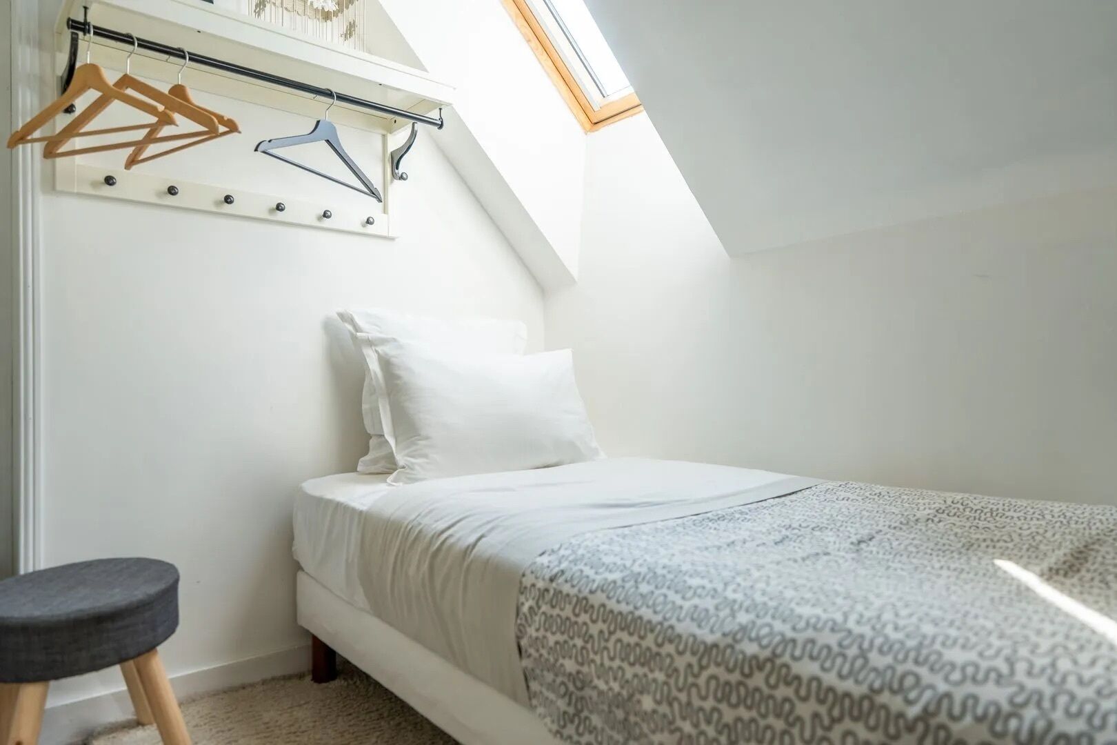 A bedroom with a bed, a hanging clothes rack, a small stool, and a skylight.