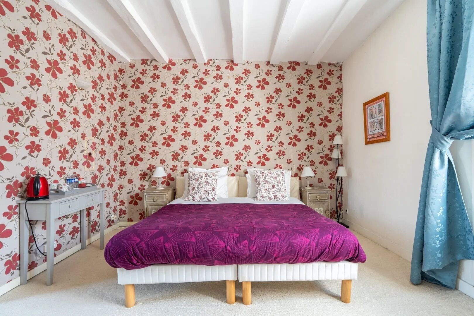 Ein Schlafzimmer mit blumigem Wandpapier, einem Bett mit lila Bettdecke, Nachttischen mit Lampen und einem blauen Vorhang.