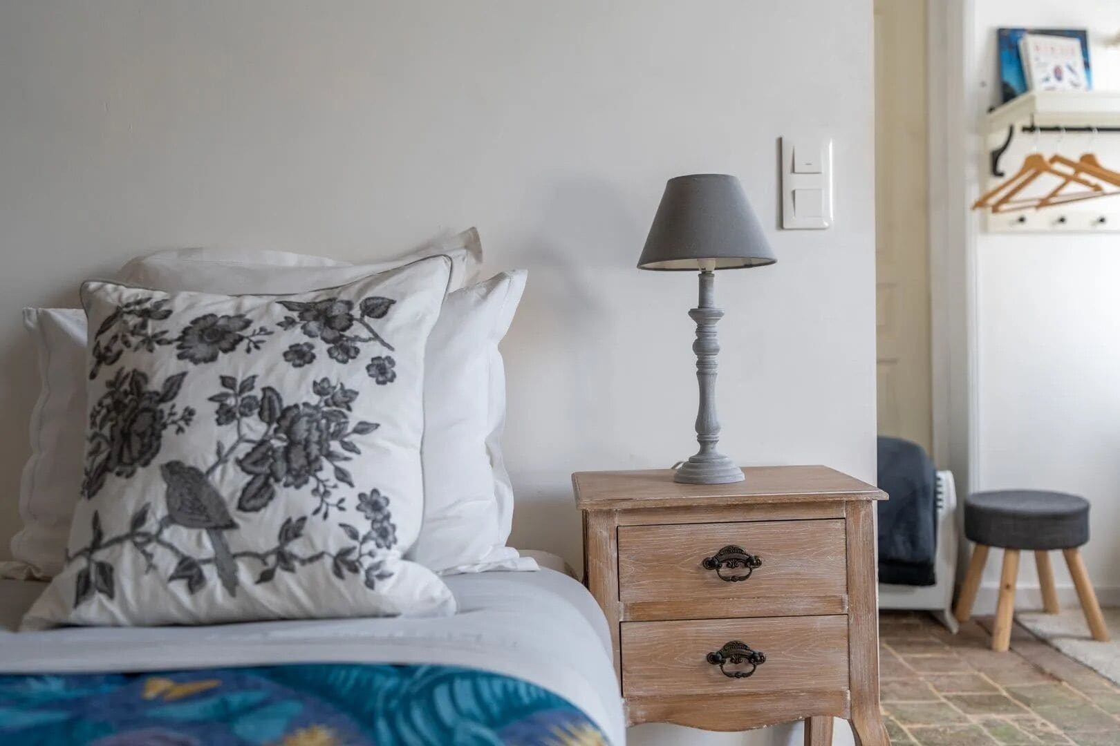 Una cama bien hecha con almohadas florales, una mesita de noche con una lámpara y un taburete de madera.