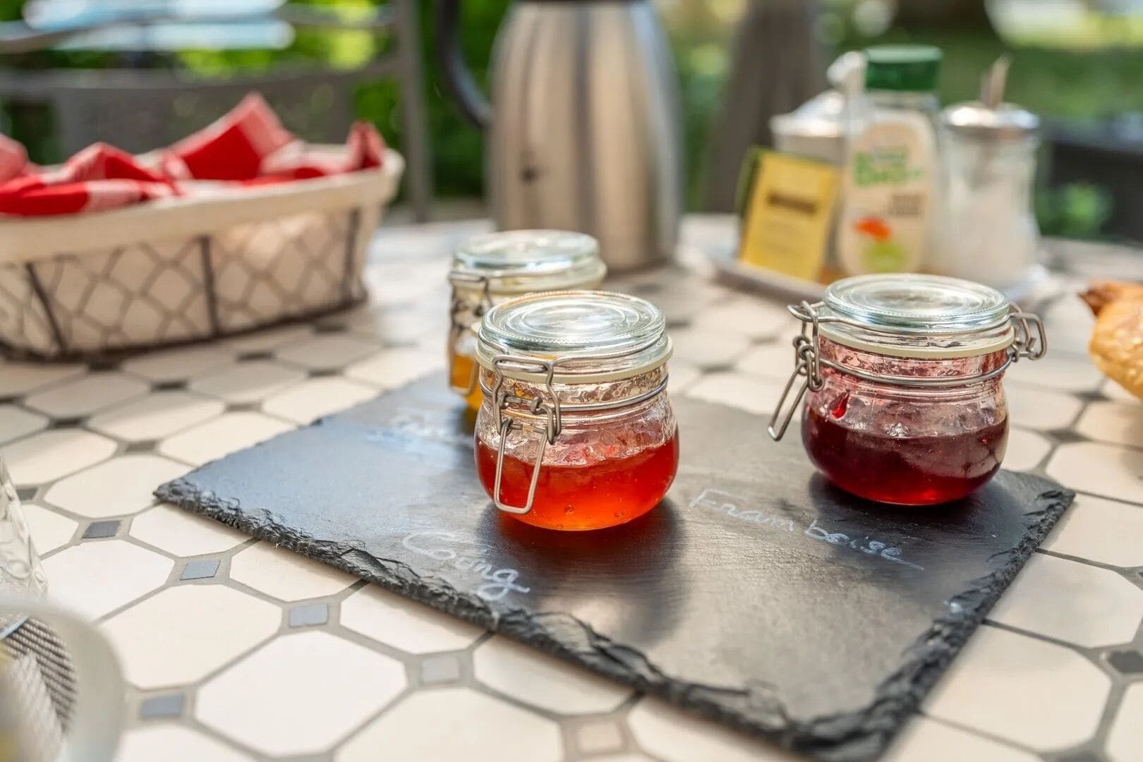 Two glass jars with metal clasps on a hexagonal tile table, a metal pitcher, and a basket of red napkins.