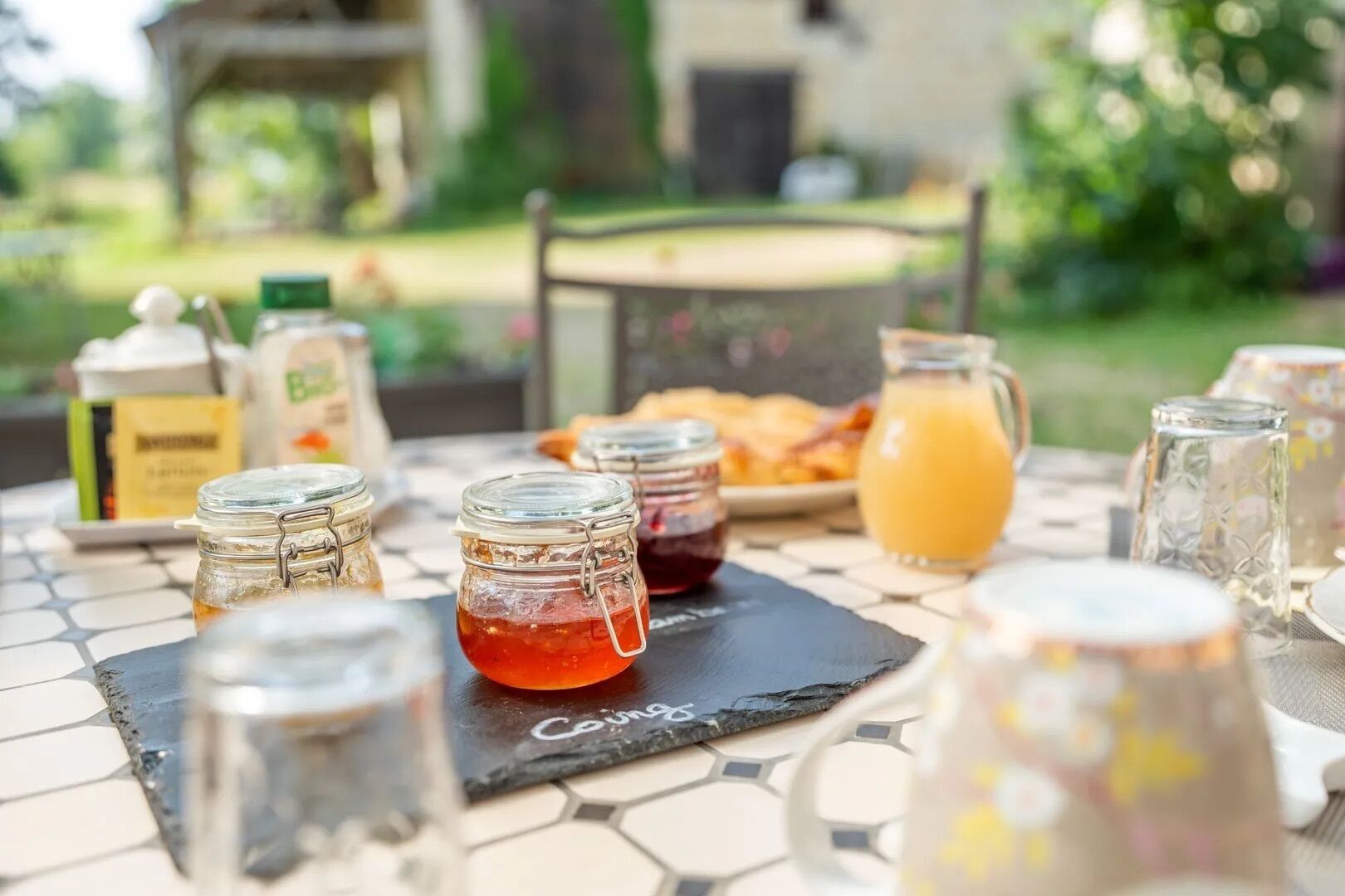 A breakfast table with jars of jam and a pitcher of orange juice, set on a tiled patio with a garden view.