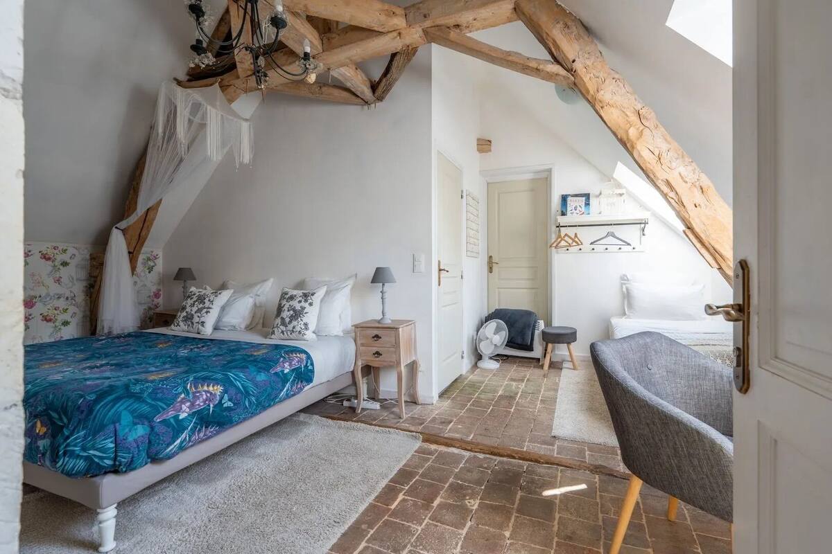 A bedroom with a sloped ceiling, a bed with a floral bedspread, a bedside table with a lamp, a chair, and a fan.