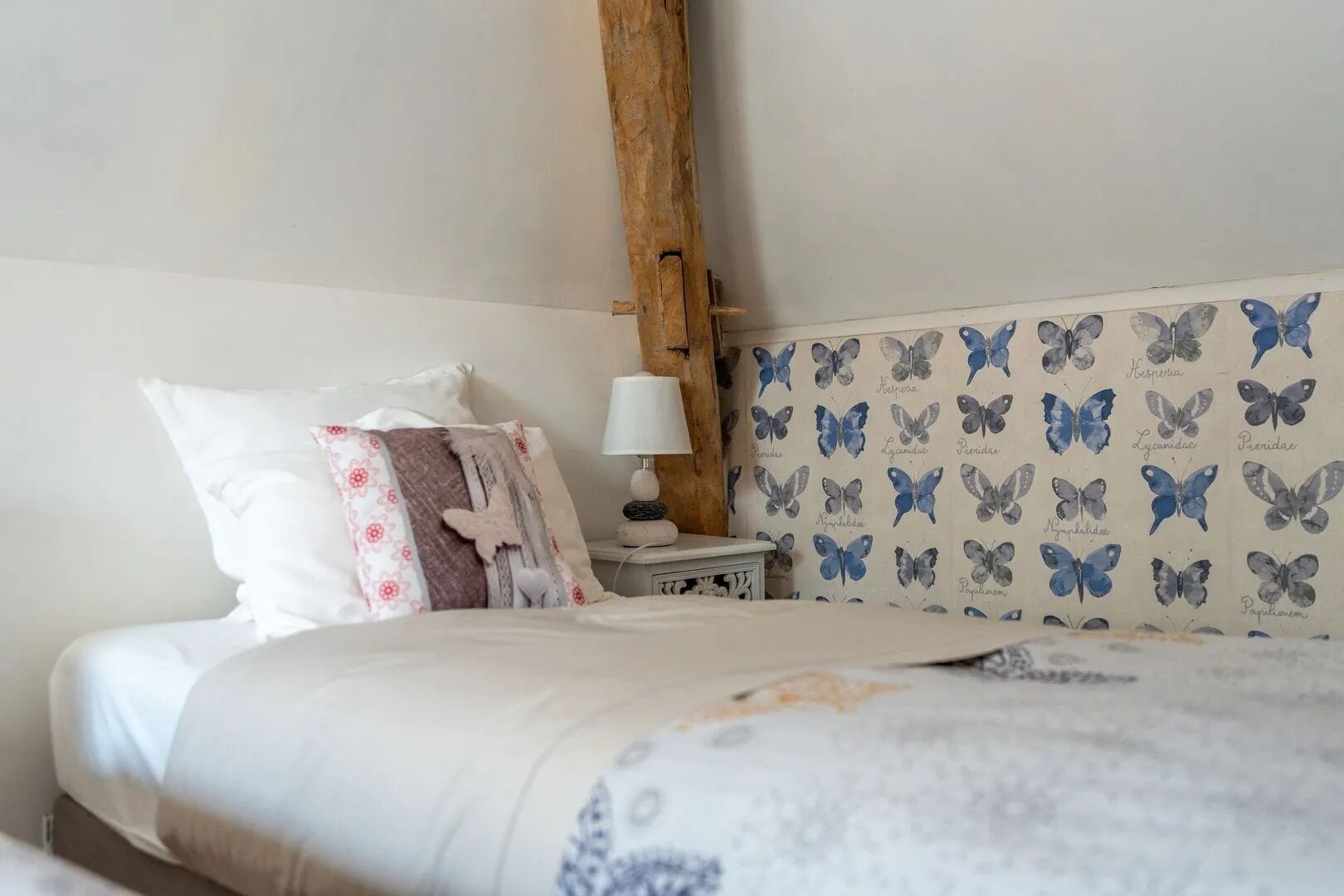 A bedroom with a bed, pillows, a nightstand, a lamp, and a wall with butterfly wallpaper.