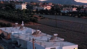 A modern building with multiple levels and a flat roof, surrounded by a landscaped area and a road. - KalliGeoM (Hersonissos)