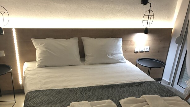 A neatly made bed with white linens, a grey quilt, and towels. There are two bedside tables, one with a lamp and the other with a small round table. - KalliGeoM (Hersonissos)