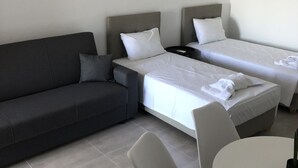 A modern hotel room with a dark grey sofa, two single beds, a small round table, and a grey armchair. - KalliGeoM (Hersonissos)
