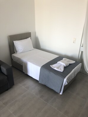 Una cama individual con cabecera gris y ropa de cama blanca, una manta gris y una toalla blanca doblada sobre la cama.