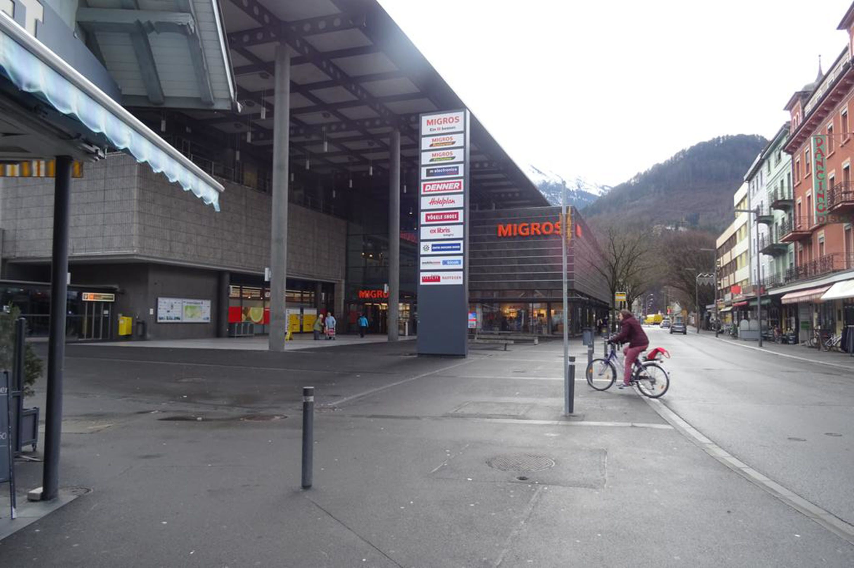 A street with a large signpost listing various businesses, including Migros.