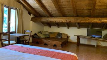 Ein gemütliches Zimmer mit Holzboden, einem Bett mit roter und weißer Bettdecke, einer Holzbank mit Kissen, einem kleinen Tisch mit Fernseher und einem Fenster mit Vorhängen.