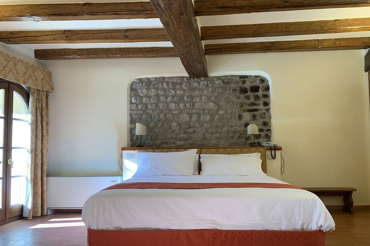 A bedroom with a stone headboard, a bed with white and orange bedding, a bedside lamp, and a wooden bench.