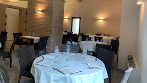 A restaurant with round tables, white tablecloths, and black chairs. There are wine glasses and cutlery on the tables. The room has a brick column and exposed beams on the ceiling. - Palazzo Dè Rossi Hotel (Sasso Marconi)