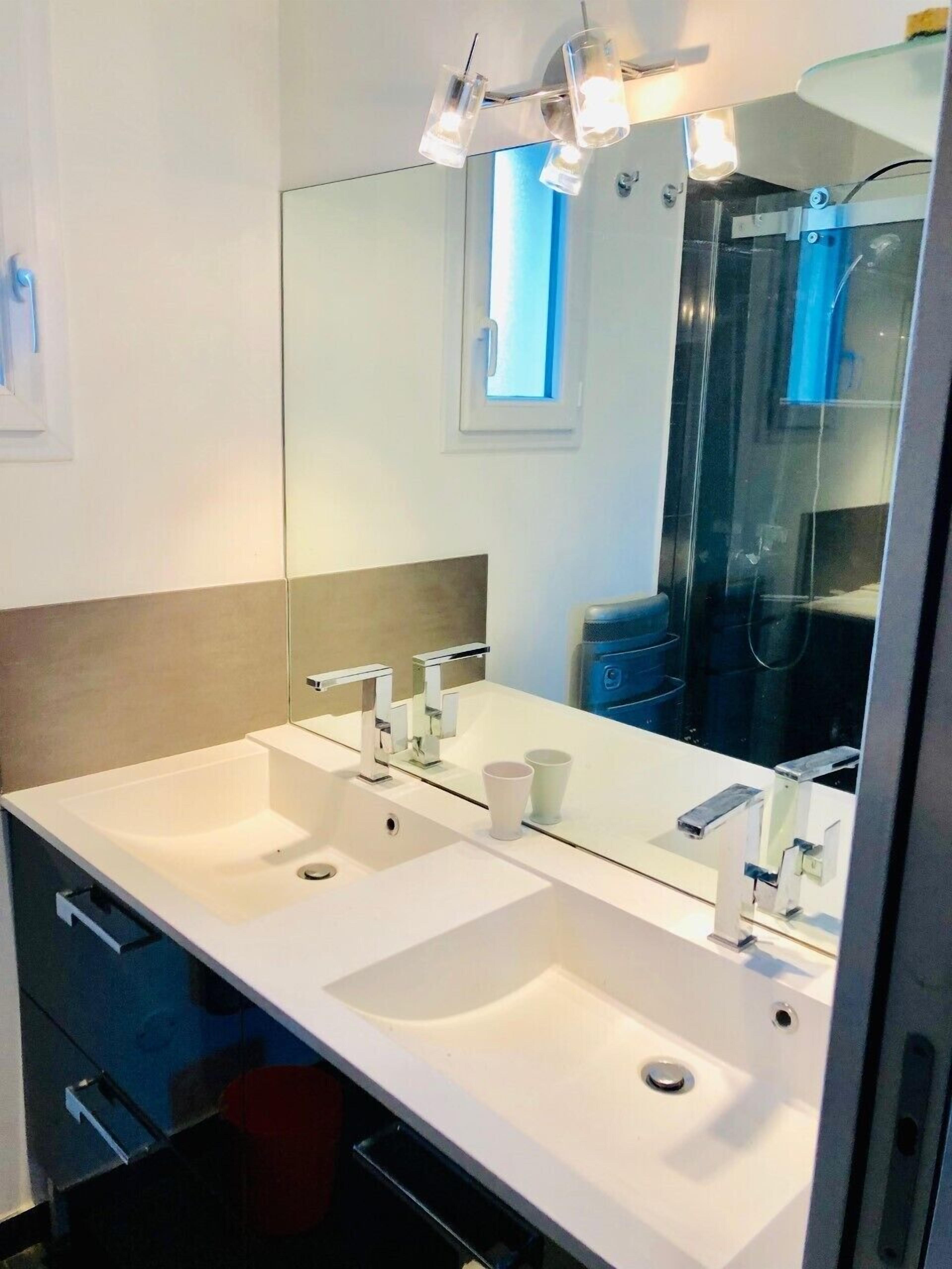 A modern bathroom with two sinks, a large mirror, and a shower visible in the reflection.