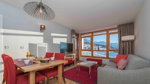 Una sala de estar moderna con una mesa de comedor de madera, sillas rojas, un sofá gris y un amplio ventanal con vistas a la montaña.