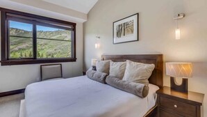 A bedroom with a large bed, a nightstand with a lamp, a framed picture on the wall, and a window with a view of hills.