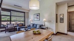 A modern living room with a wooden dining table, a blue sofa, and a large window offering a view of the outdoors.