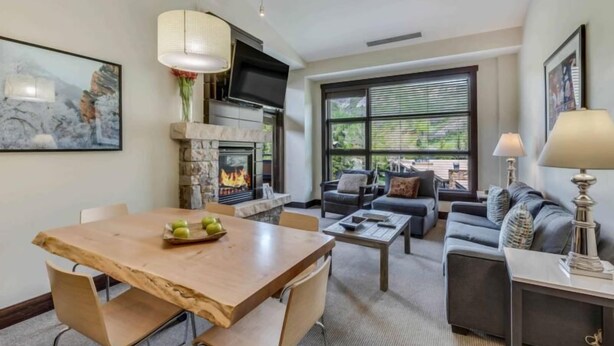 A modern living room with a stone fireplace, a dining table with apples, a sofa, and a TV mounted on the wall.