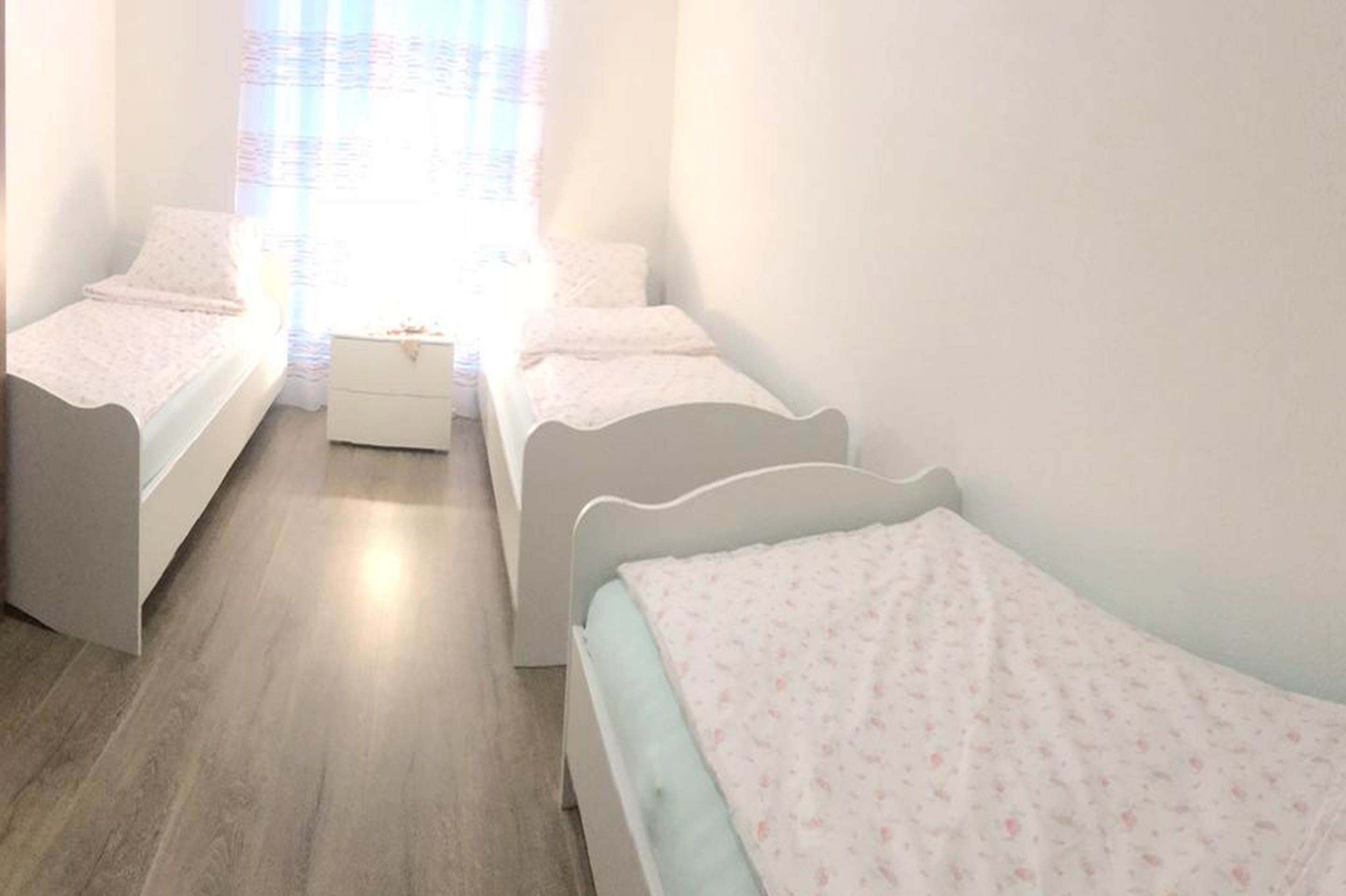 Ein Zimmer mit zwei Einzelbetten, einer kleinen weißen Kommode und einem Fenster mit durchsichtigen Vorhängen.