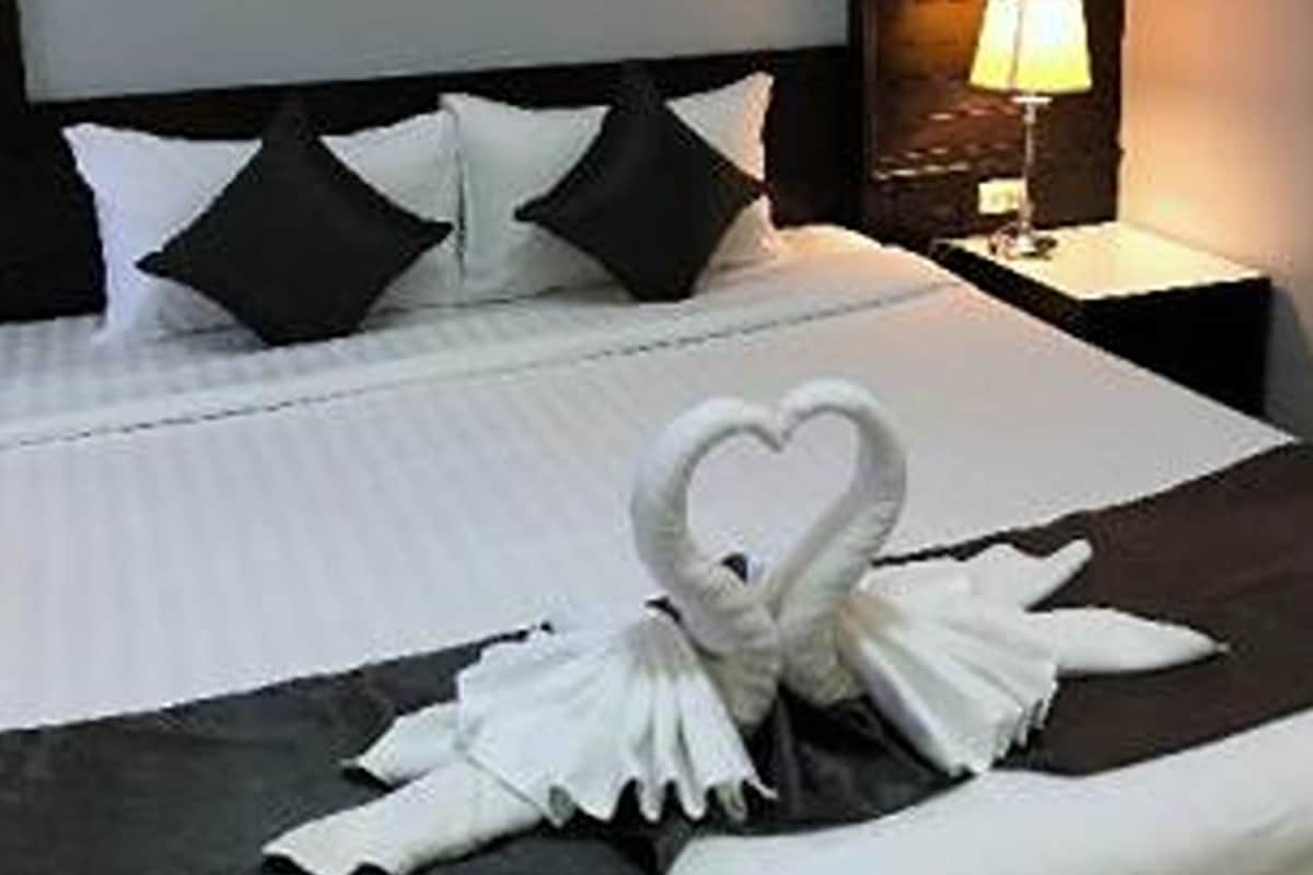 A neatly made bed with a heart-shaped towel arrangement, a lamp on the side table, and a dark headboard.