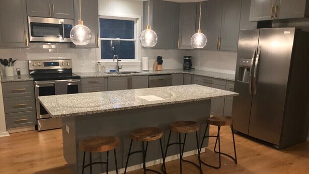 A modern kitchen with grey cabinets, stainless steel appliances, a granite island with bar stools, and pendant lighting.