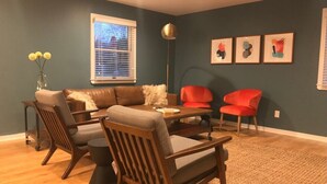A living room with a sectional sofa, two red chairs, a coffee table, and a floor lamp. There is a window with blinds, a vase with flowers, and three framed pictures on the wall.