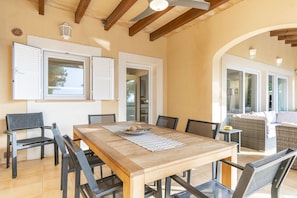 A dining area with a wooden table and chairs, a view of the outdoors, and a wicker sofa. - EMBAT - Seafront house with garden, pool and bbq. (Colonia San Pedro)
