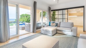 A modern living room with a grey sofa, a white ottoman, and a large window offering a view of a pool and greenery. - EMBAT - Seafront house with garden, pool and bbq. (Colonia San Pedro)