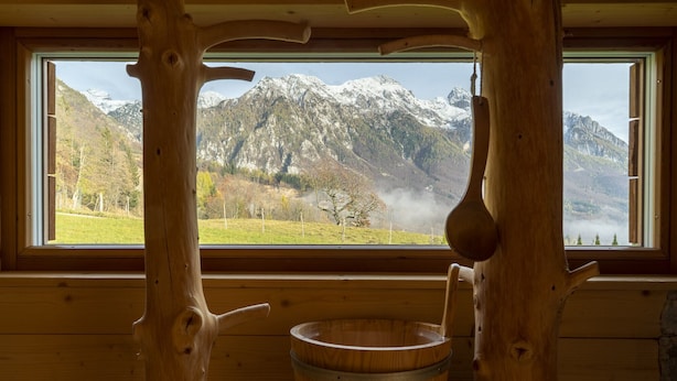 Una cabaña de madera con un amplio ventanal que ofrece vistas a montañas nevadas y un valle verde.