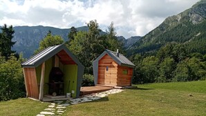 Dos cabañas de madera pequeñas con techos a dos aguas, un sendero de piedra que las conduce, y un telón de montañas de fondo.