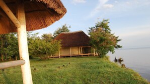 A thatched-roof hut with a wooden deck, surrounded by grass and trees, near a body of water. - Ntoroko Game Lodge (Ntoroko)