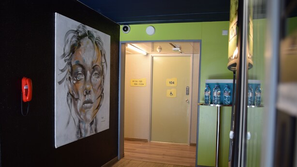 A hallway with a painting on the wall, a door with a wheelchair accessibility sign, and a water dispenser. - Hotel Design Booking (Villeneuve-le-Roi)