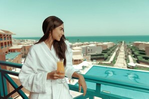Una persona vestida con una bata blanca sosteniendo una bebida, con vistas a un complejo turístico con piscina y edificios al fondo.