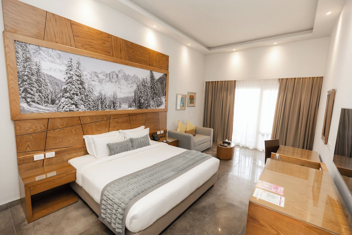 Moderni hotellihuone, jossa on suuri sänky, sohva, televisio ja puinen pääty, jossa on maalattu kuva vuoristomaisemasta.