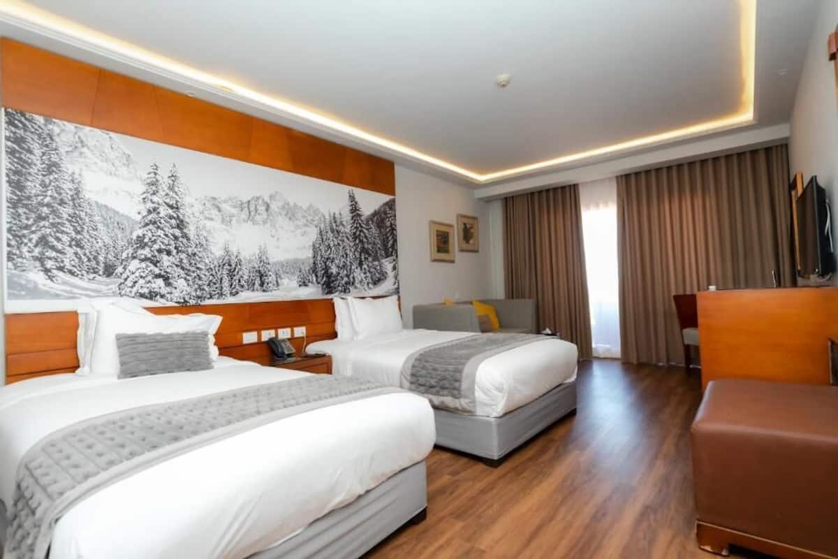 Hotellihuone, jossa on kaksi sänkyä, puinen pääty, suuri seinälle ripustettu taulu, ruskea nahkainen nojatuoli ja ikkuna, jossa on verhot.