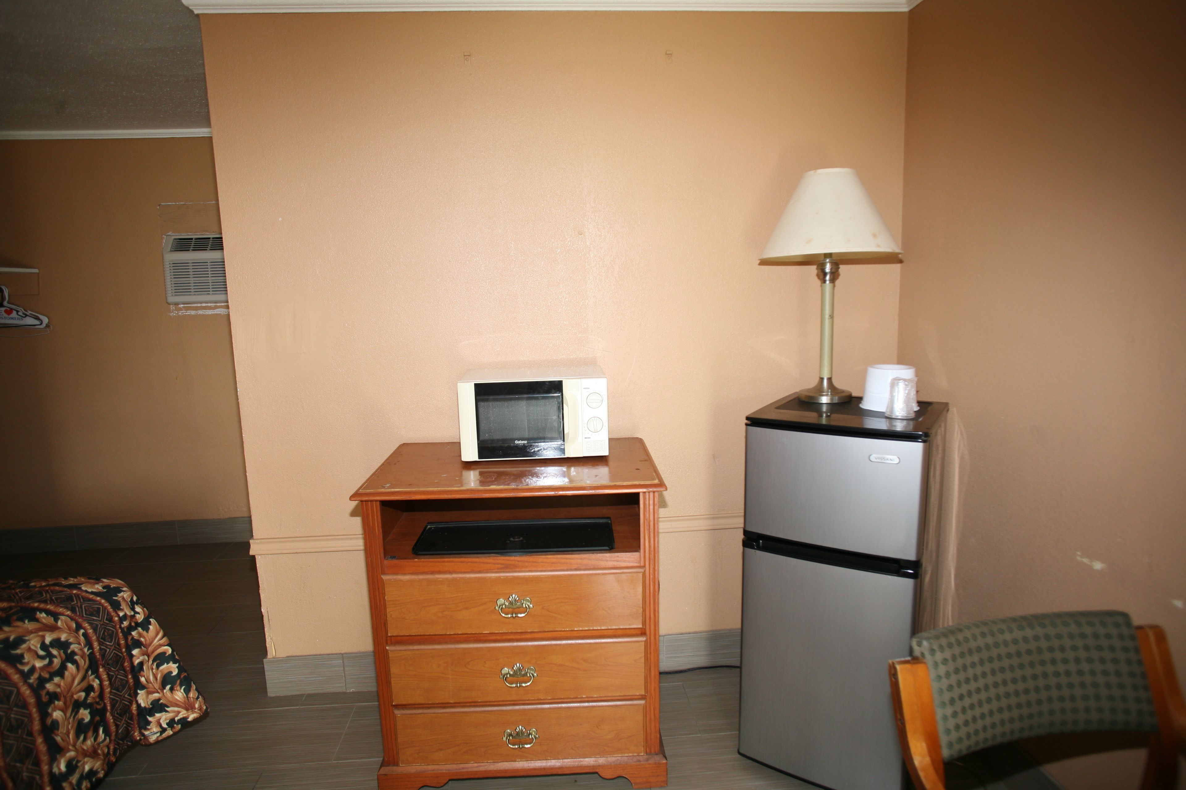 A compact room with a wooden dresser, a microwave, a refrigerator, a lamp, and a chair.