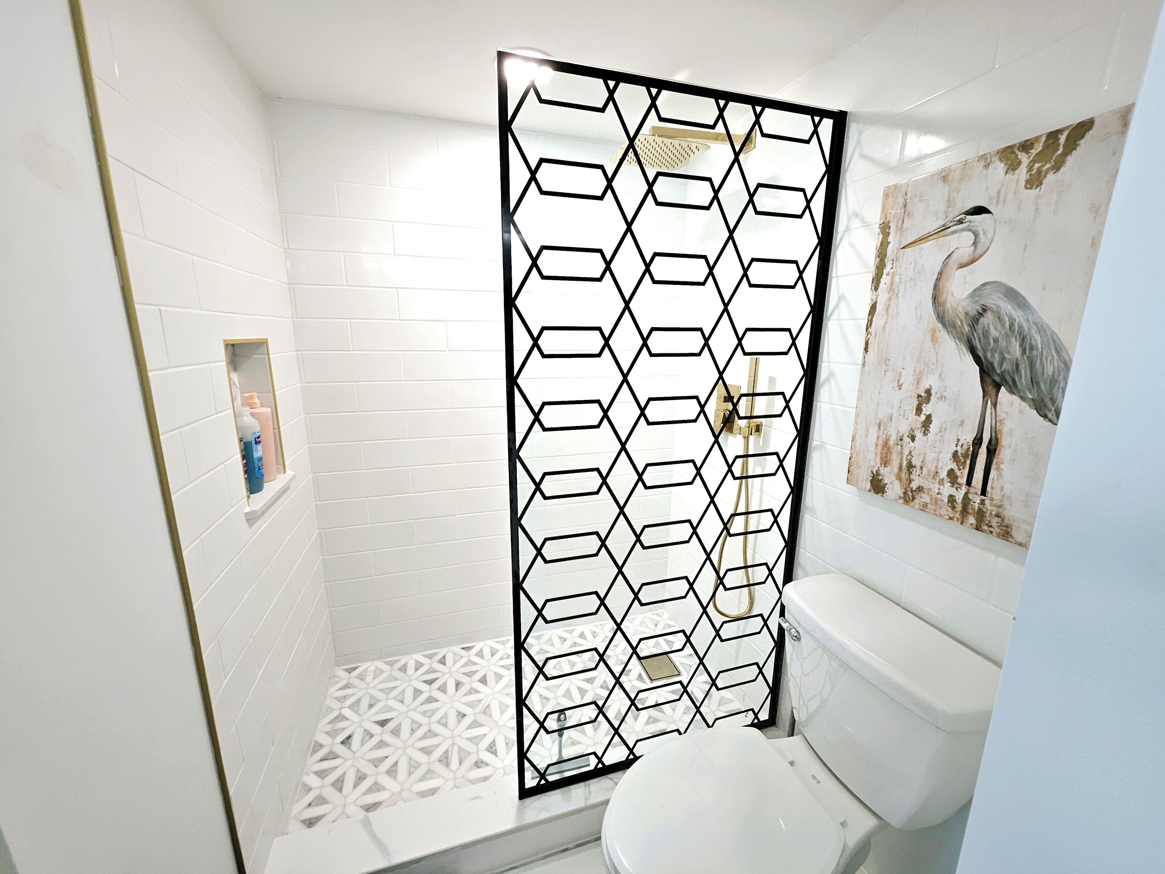 A bathroom with a white tiled floor, a white toilet, a shower with a black geometric partition, and a wall-mounted sink with a gold frame.