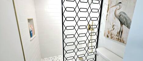 A bathroom with a white tiled floor, a white toilet, a shower with a black geometric partition, and a wall-mounted sink with a gold frame.