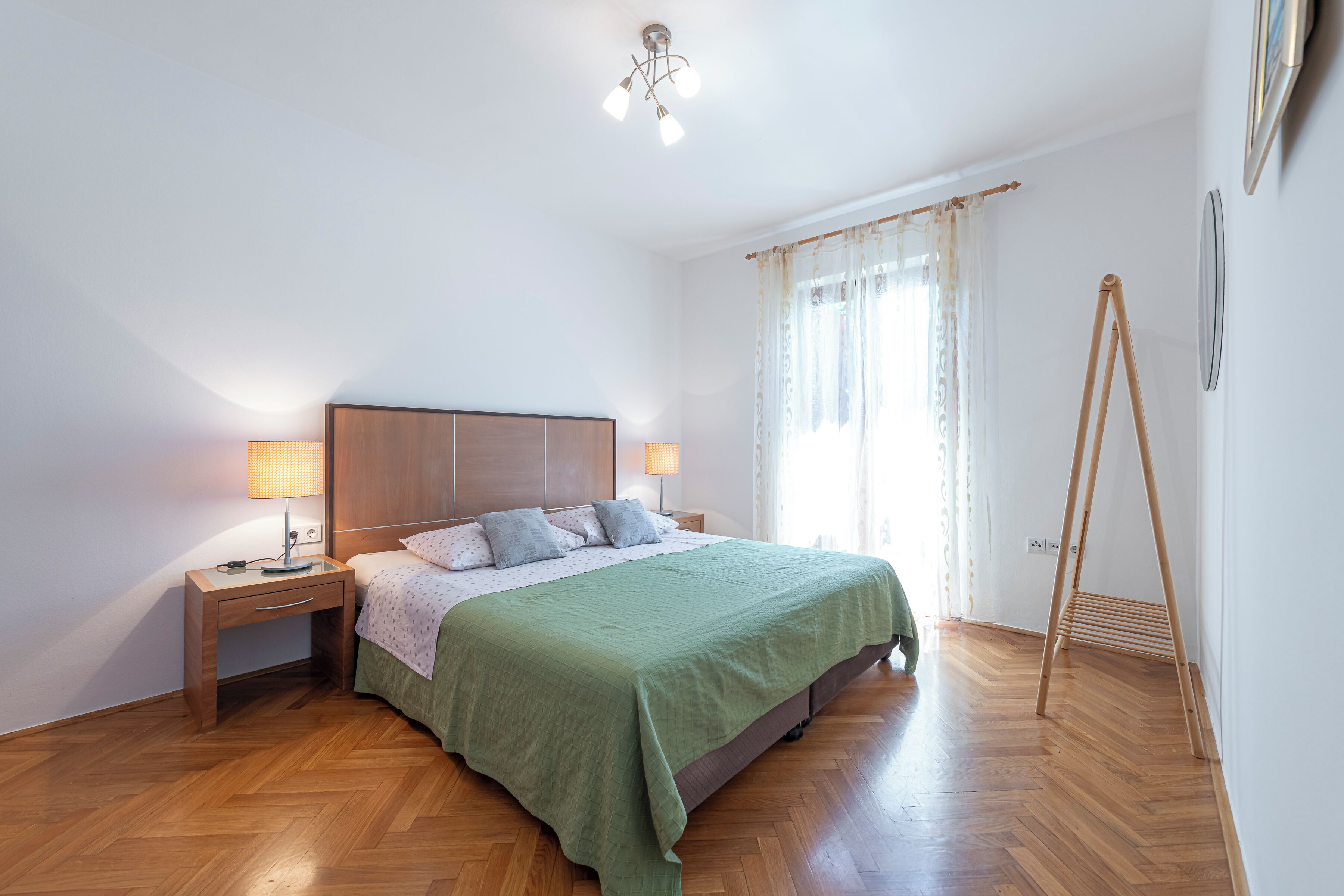 Un dormitorio con cama, mesitas de noche, lámpara, ventana con cortinas y suelo de madera.