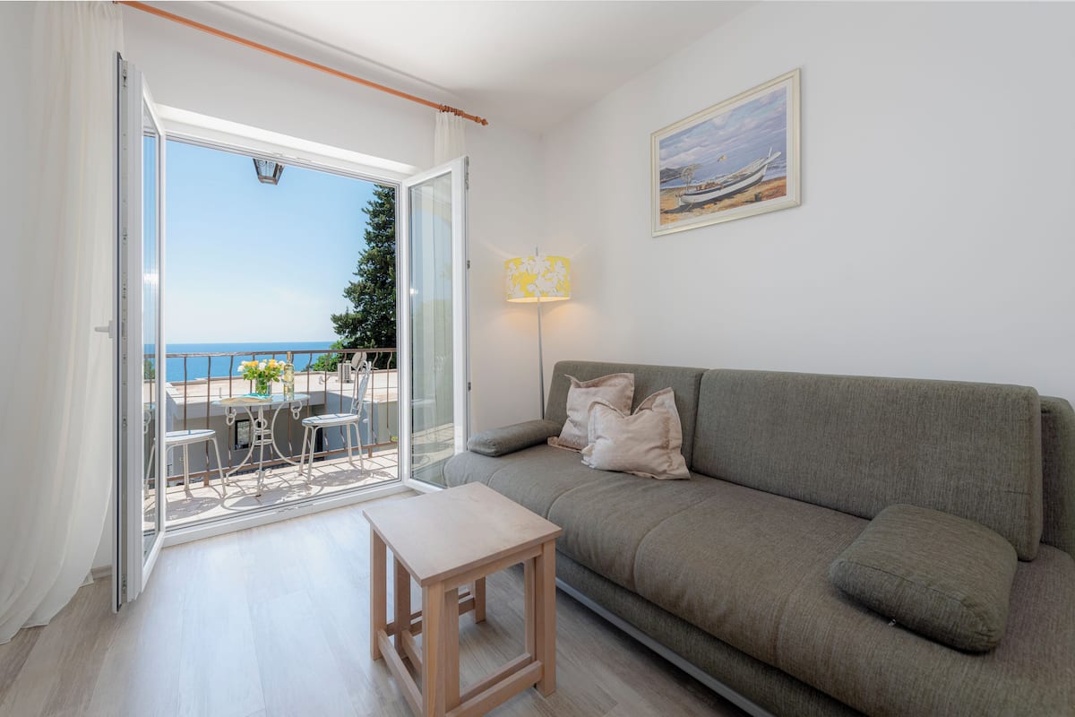 Un salon moderne avec un canapé gris, une petite table en bois et une porte en verre donnant sur un balcon avec vue sur la mer.