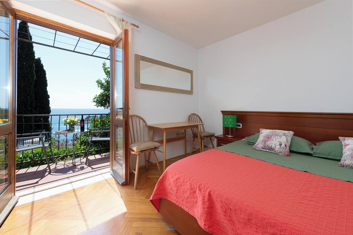Une chambre avec un balcon, un lit avec des coussins verts, un bureau en bois et une chaise.