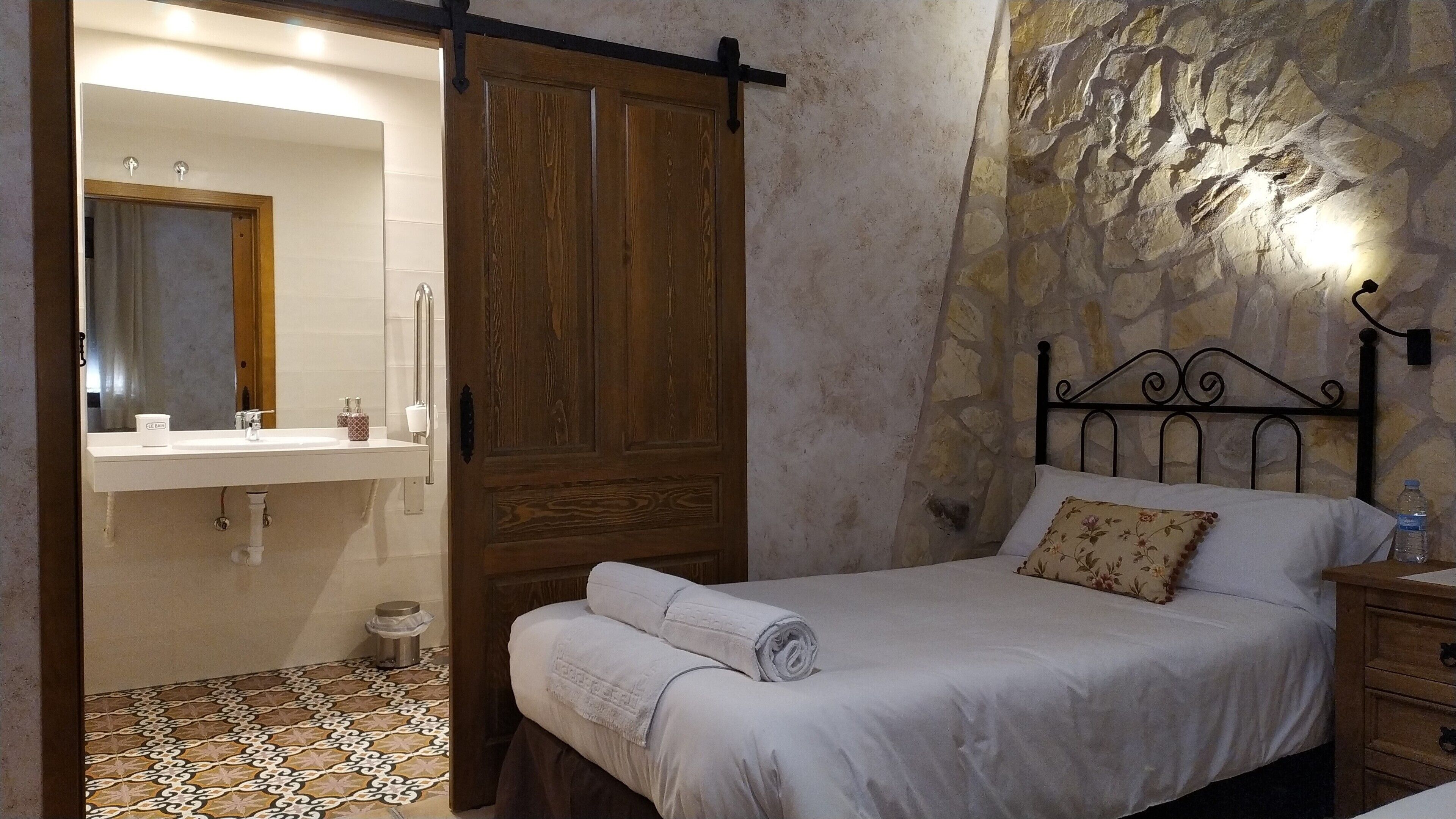 Un dormitorio con cama, mesita de noche, armario de madera, baño con lavamanos y espejo, y pared de piedra.