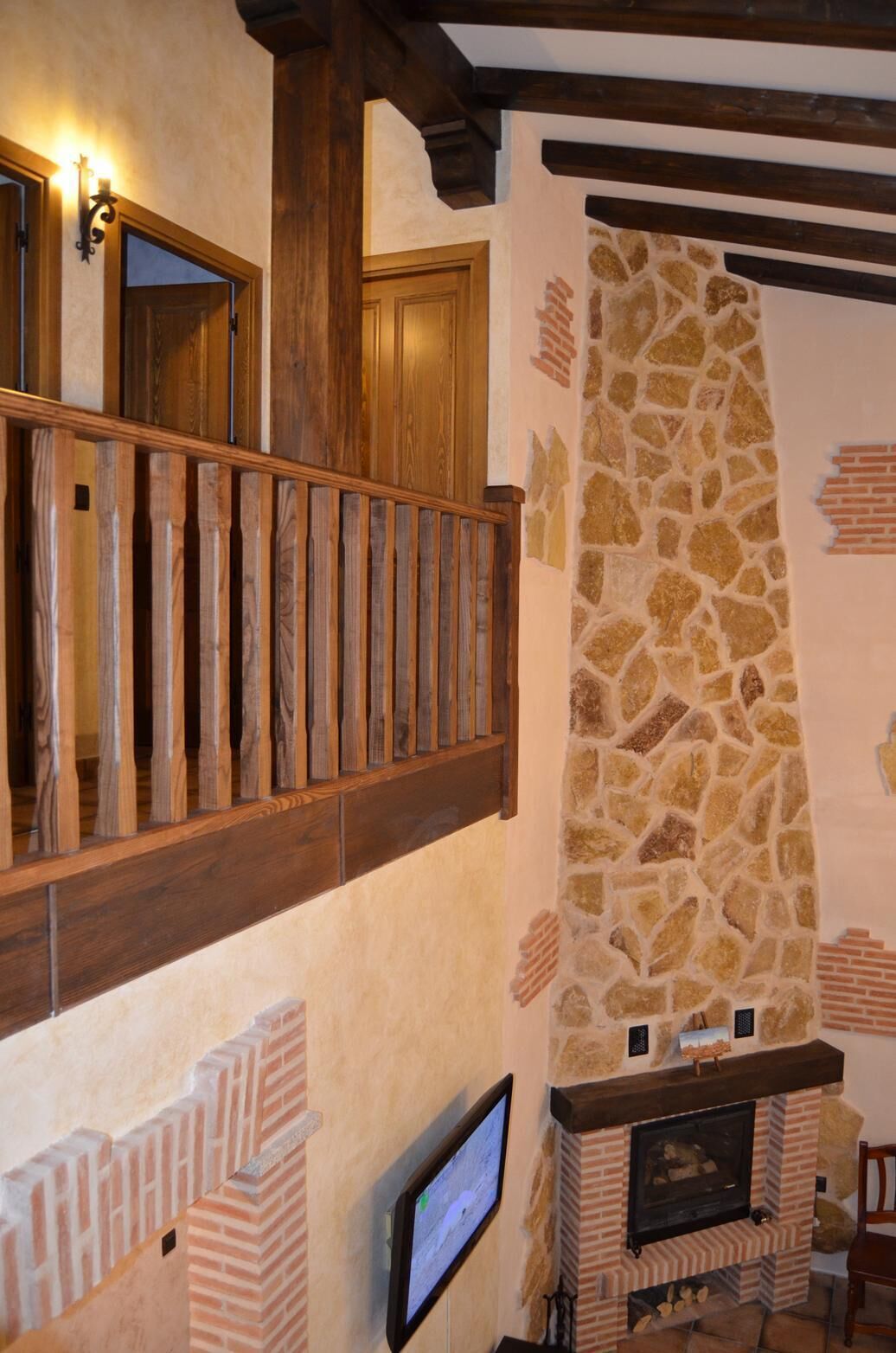 Un interior rústico con chimenea de piedra, escalera de madera y un televisor montado en la pared.