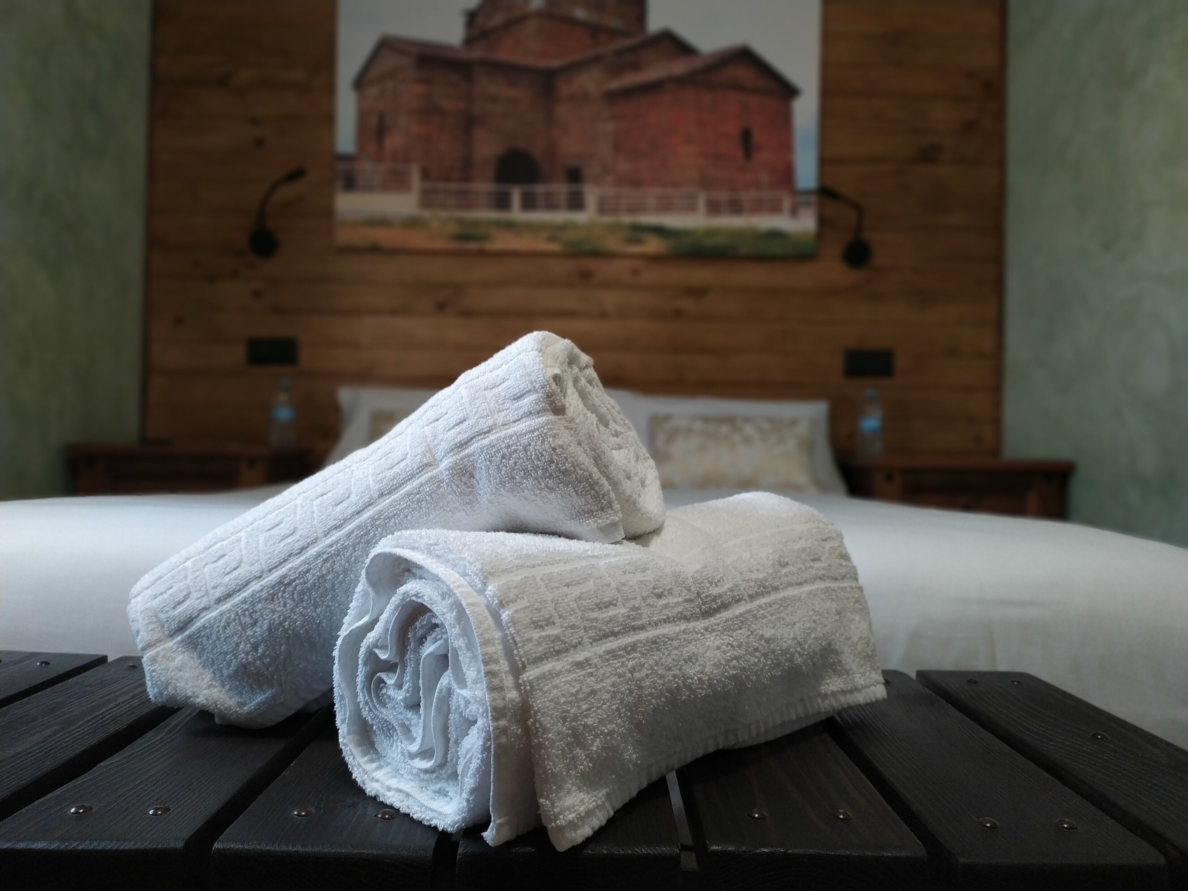 Un lit bien fait, avec du linge de lit blanc et deux serviettes blanches roulées, posées sur une table en bois dans une pièce aux murs en bois. On aperçoit, au fond, un tableau représentant un bâtiment.