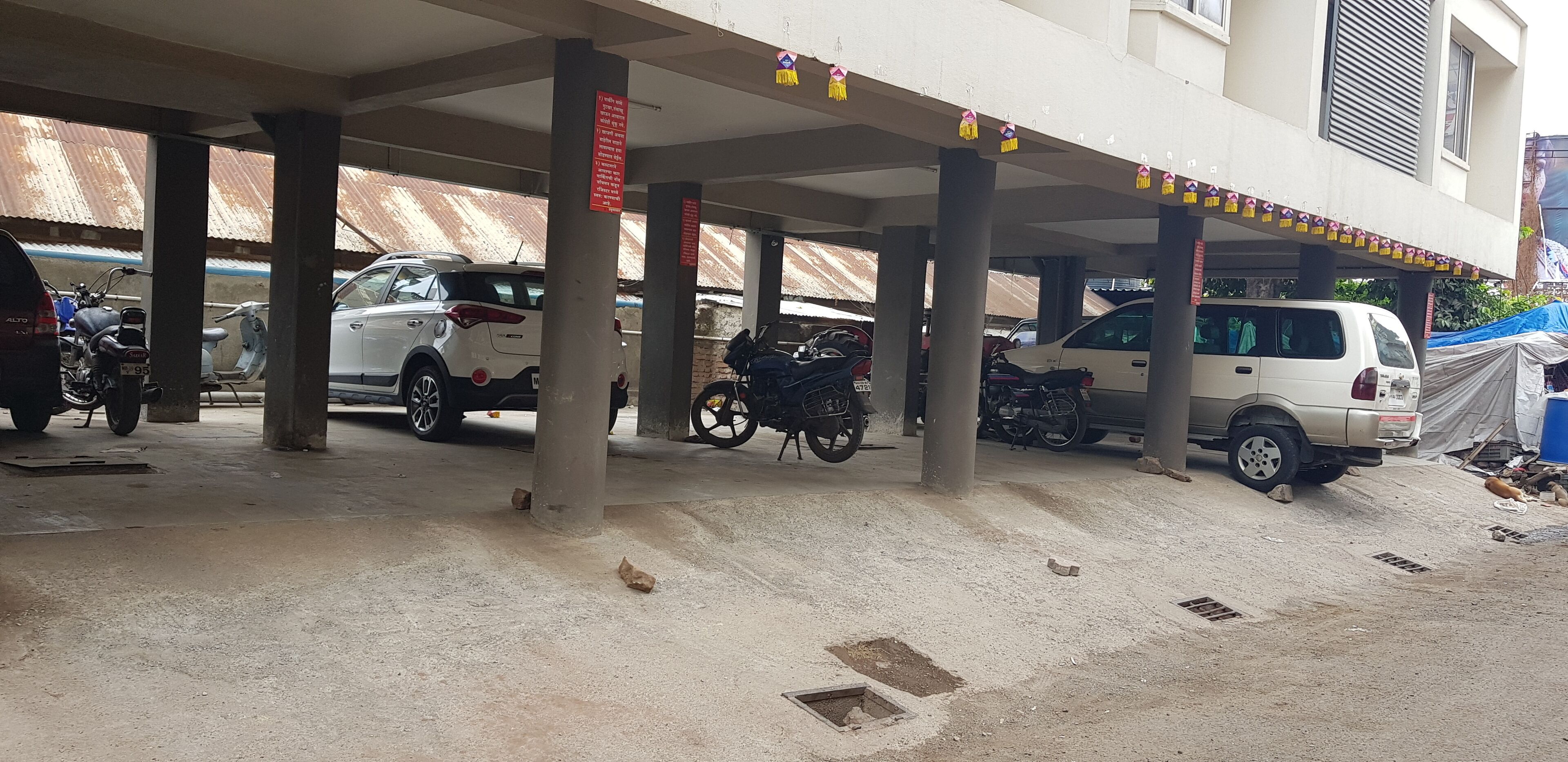 A covered parking area with multiple vehicles, including cars and motorcycles, parked under a structure with pillars and a tiled roof.