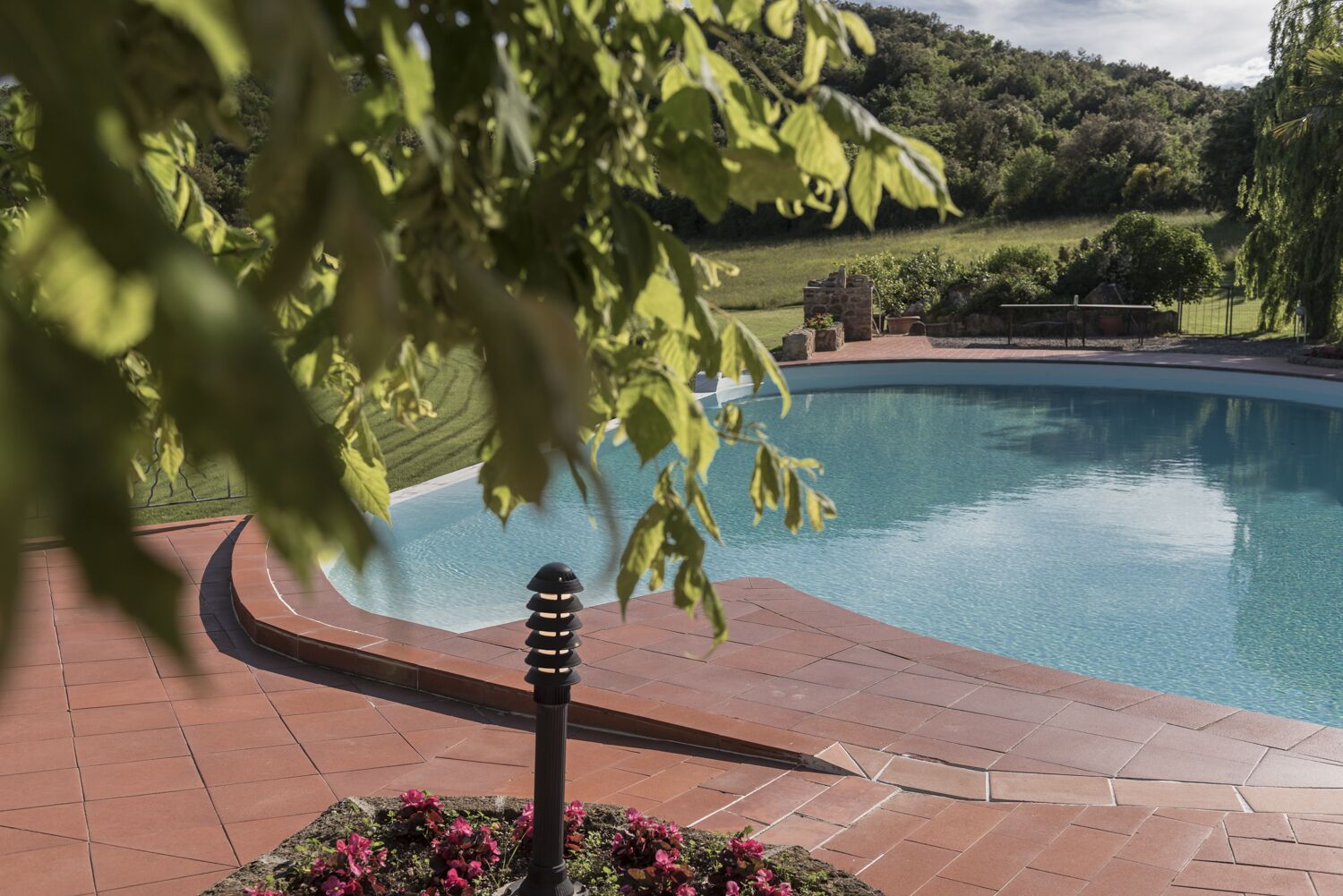 Una piscina de borde curvo, rodeada de baldosas de terracota y un macizo de flores rosadas.
