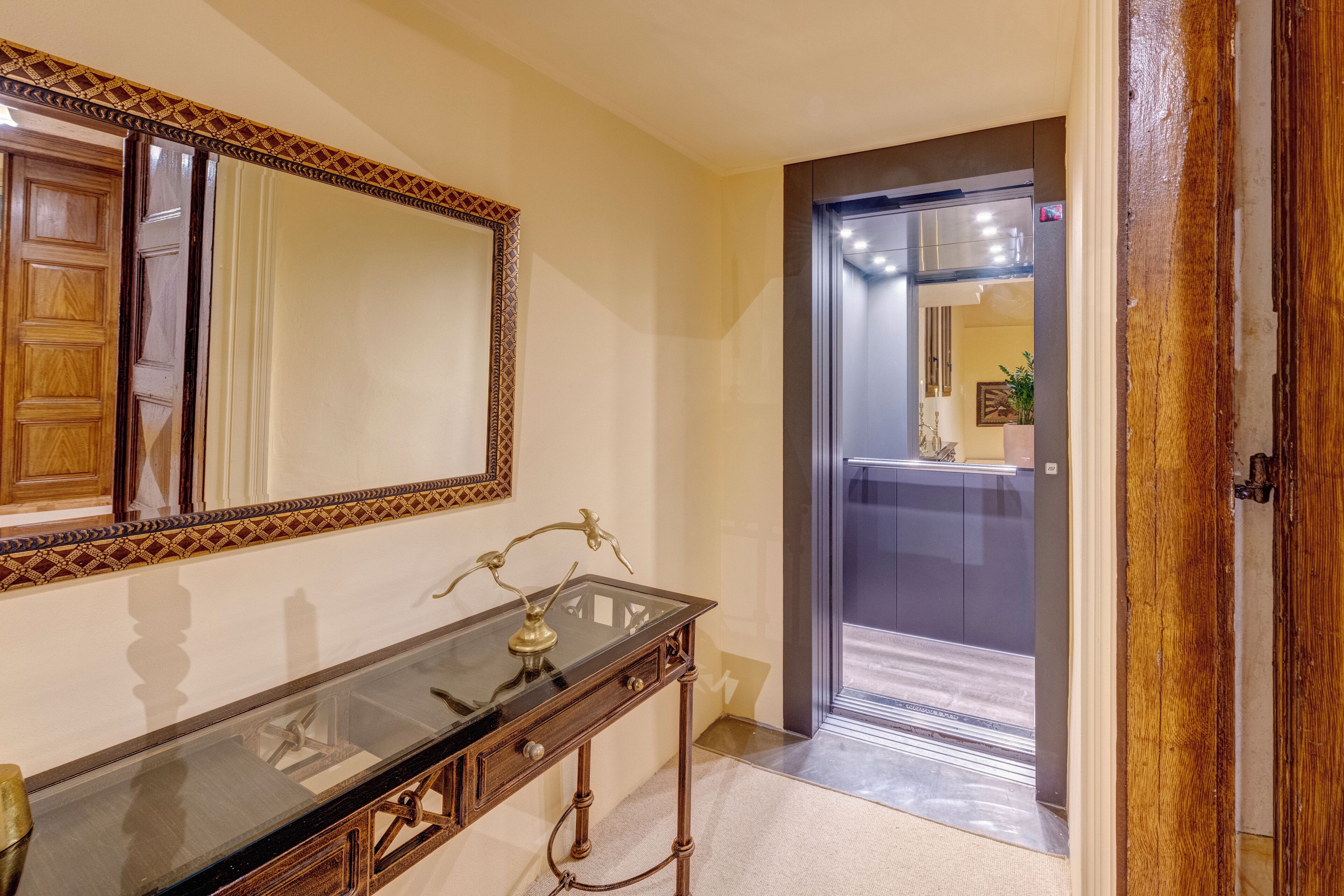 Um corredor com porta de madeira, espelho e mesa de vidro com abajur de latão.