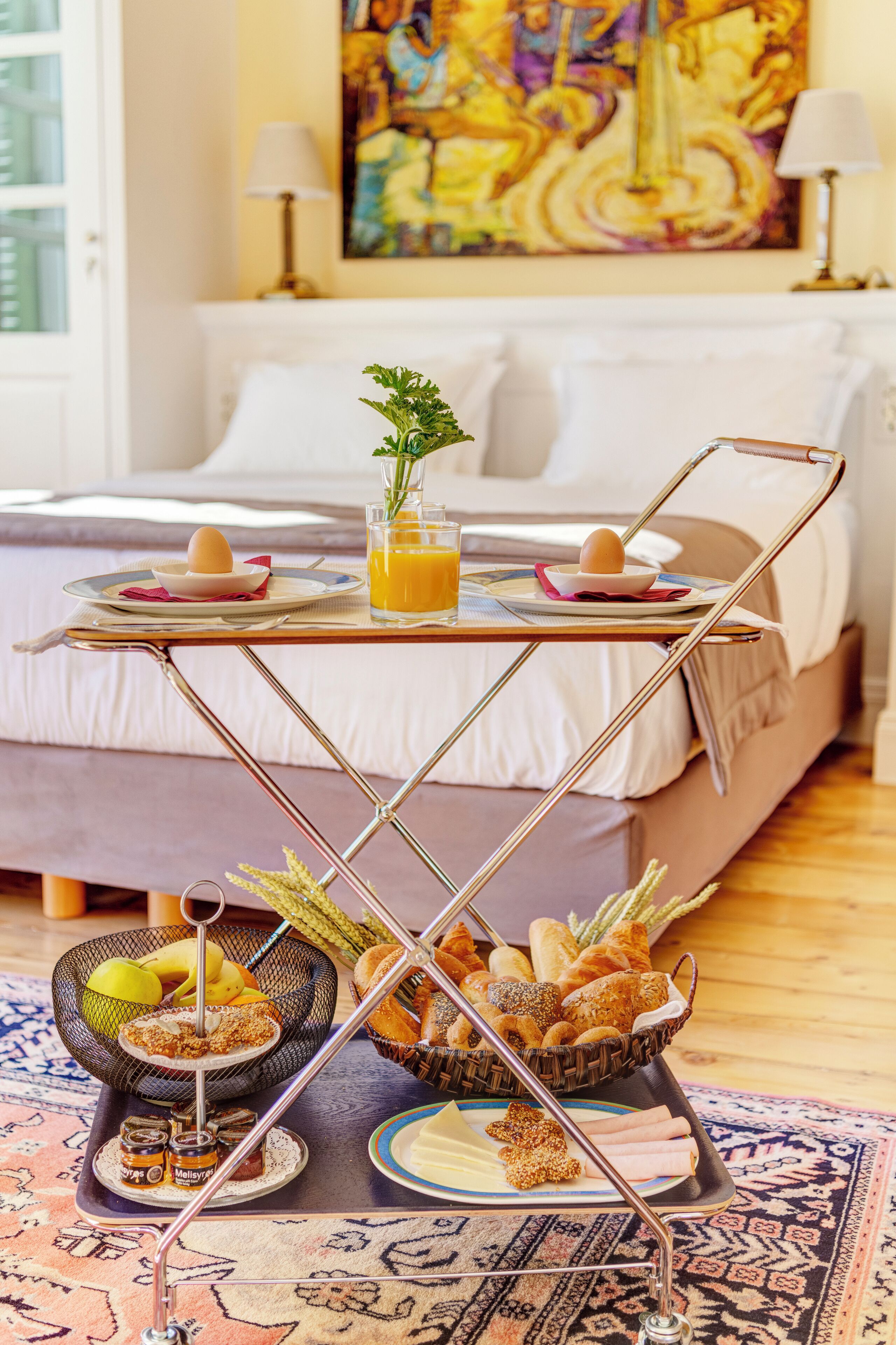 A breakfast setup with a tray of eggs, juice, and pastries on a table in front of a bed with a painting on the wall.