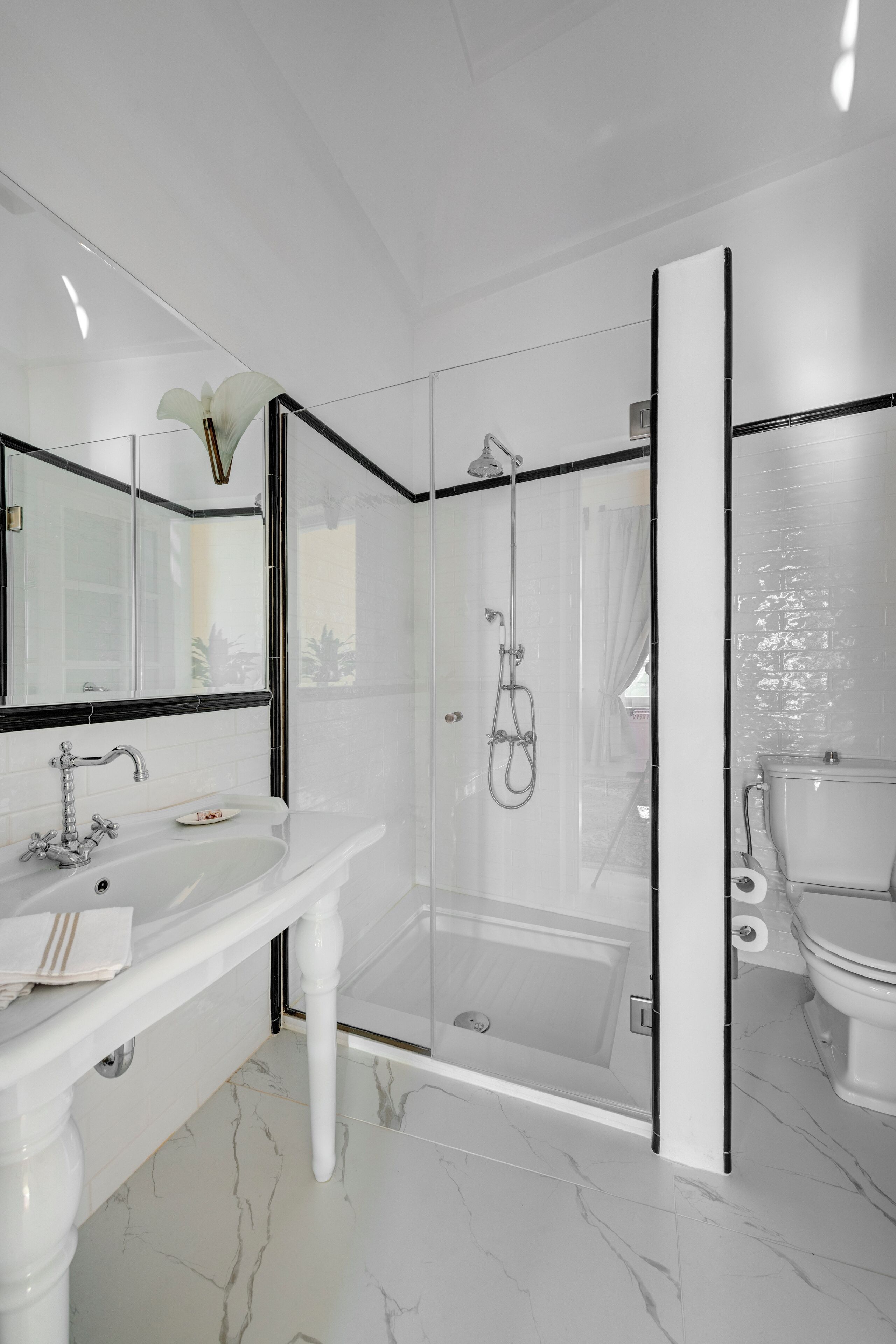 Un baño moderno con piso de mármol blanco, ducha con cerramiento de vidrio, lavamanos blanco con espejo y un inodoro.