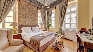A spacious bedroom with a large bed, a bedside table with a lamp, a desk with a chair, and a breakfast trolley. - Palazzo Santa Maria - Historic Hotels of Europe (Syros)
